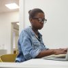 Serious African woman in eyeglasses sitting at the table and typing on laptop computer at office