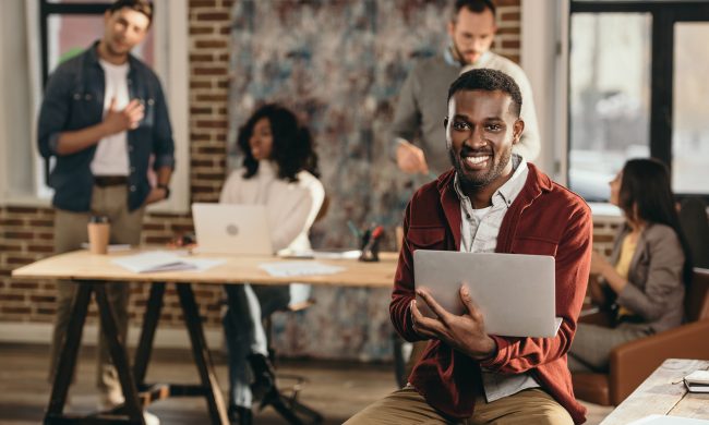 african american casual businessman sitting with laptop and colleagues working behind in loft office