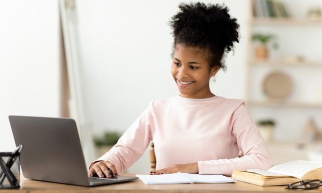 African American Teenager Girl Doing Homework On Laptop Computer Sitting At Table Indoors. Selective Focus