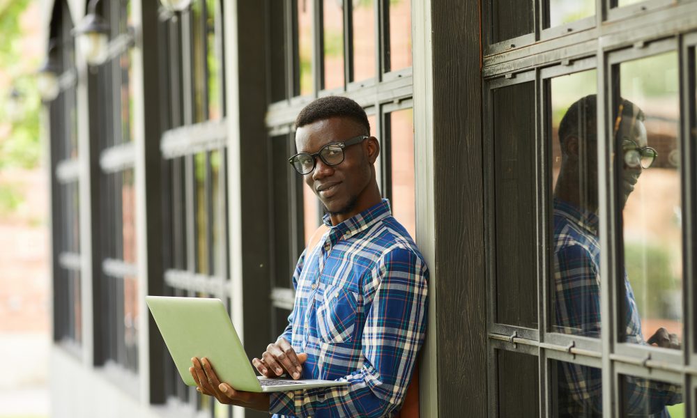 Waist up portrait of smart African student holding laptop posing outdoors in college campus, copy space