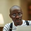 Head and shoulders portrait of contemporary African student wearing glasses using laptop while studying in library, copy space